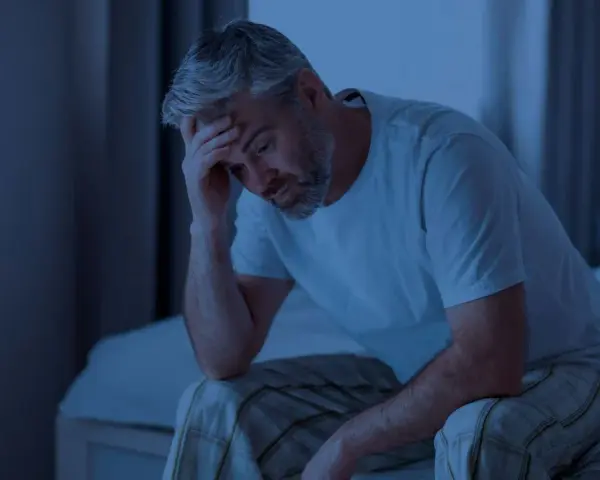 Ein Mann sitzt im Dunkeln auf seinem Bett, die Hand an der Stirn. Er fragt sich, wie lange man ohne Schlaf auskommt.
