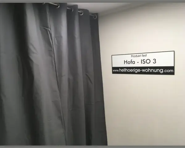 Grauer Schallschutzvorhang im Test. Ein Schild mit "Produkt-Test Hofa - ISO 3" und "www.hellhoerige-wohnung.com" hängt an der Wand.