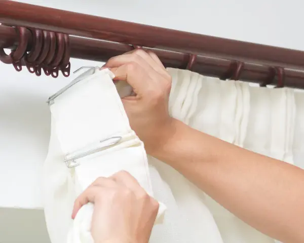 Hände befestigen weiße Gardinen an einer dunklen Holzstange mit Ringen.