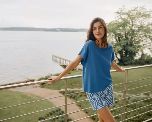 Frau auf Balkon mit Blick auf See, trägt blaues Oberteil und gemusterte Shorts. In Modal schwitzt man nicht, es ist angenehm kühl.