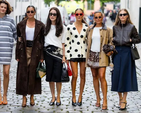 Sechs Frauen in stylischen Outfits, darunter ein gestreiftes Kleid und ein Blazer mit nordischen Mustern, posieren auf einer Kopfsteinpflasterstraße.