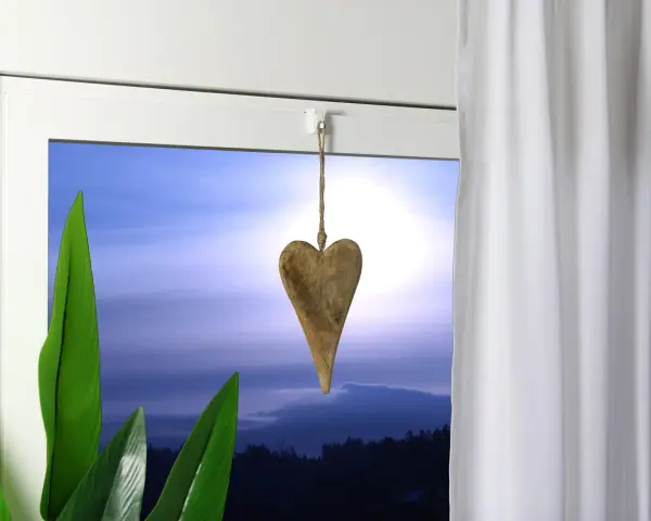 Holzherz hängt an einer Schnur, um die Gardinenstange zu dekorieren. Grüne Blätter und ein dramatischer Himmel im Hintergrund.