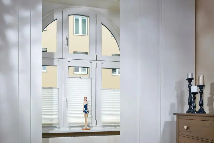 Weiße Schiebegardinen in alten Schienen vor einem Bogenfenster mit Blick auf ein Gebäude. Eine Puppe steht auf der Fensterbank.