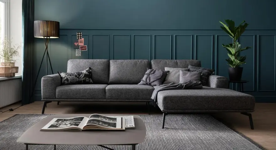 Ein moderner Wohnbereich mit einem anthrazitfarbenen Sofa, das auf einem gemusterten Teppich steht. Ein Couchtisch mit einem offenen Buch und eine Stehlampe vervollständigen das Bild.