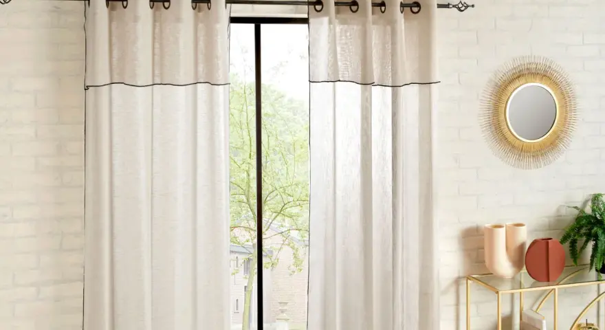 Helle Gardinen mit schwarzer Umrandung sind hochgesteckt und rahmen ein Fenster mit Blick ins Grüne ein.