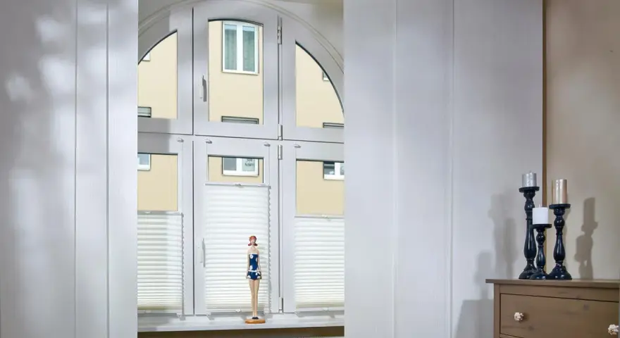 Weiße Schiebegardinen in alten Schienen vor einem Bogenfenster mit Blick auf ein Gebäude. Eine Puppe steht auf der Fensterbank.