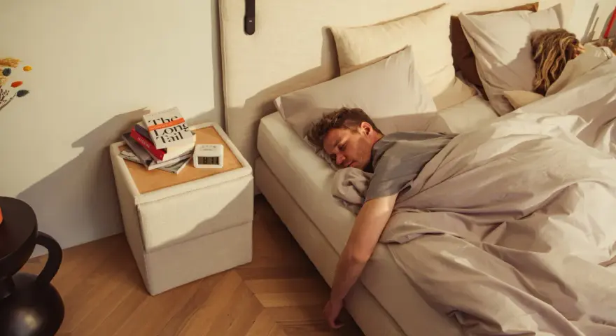Mann schläft tief und fest auf einem Bett, das sich wie **Kaltschaum** anfühlt. Neben dem Bett liegen Bücher und ein Wecker.