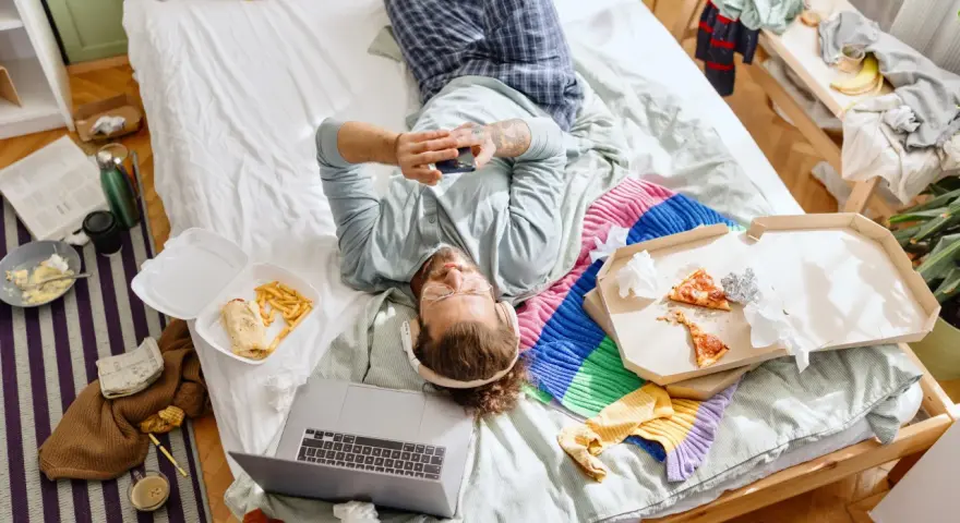 Mann liegt im Chaos auf dem Bett, umgeben von Essensresten und Kleidung. Ein typischer Tag des "bed rotting".