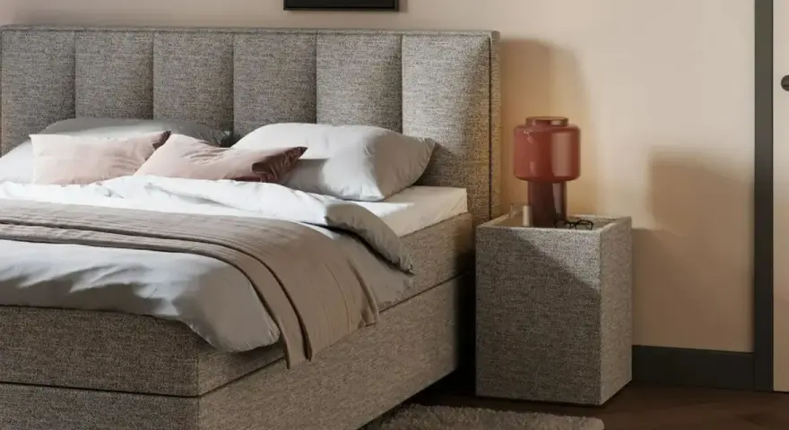 Gemütliches Queensize Bett mit grauem Stoffbezug, Kissen und Decke. Daneben ein Nachttisch mit Lampe.