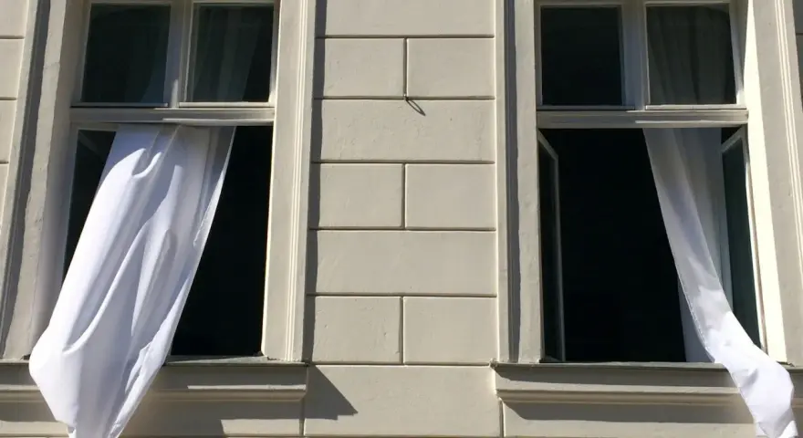 Weiße Gardinen wehen aus zwei offenen Fenstern eines hellen Gebäudes. Die Frage "Gardinen waschen wieviel Grad?" beschäftigt die Hausfrau.