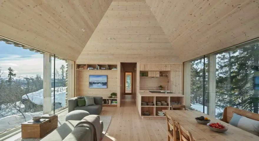 Gemütliches Wohnzimmer mit Holzwänden und Decke, die ein kariertes Muster andeuten. Große Fensterfronten bieten Blick auf verschneite Landschaft.