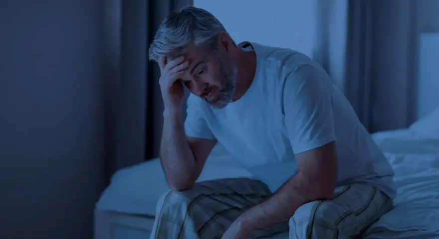 Ein Mann sitzt im Dunkeln auf seinem Bett, die Hand an der Stirn. Er fragt sich, wie lange man ohne Schlaf auskommt.