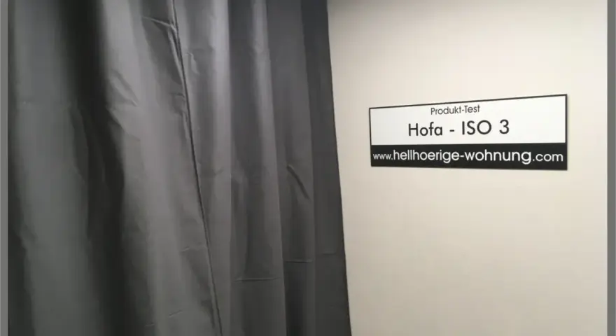 Grauer Schallschutzvorhang im Test. Ein Schild mit "Produkt-Test Hofa - ISO 3" und "www.hellhoerige-wohnung.com" hängt an der Wand.