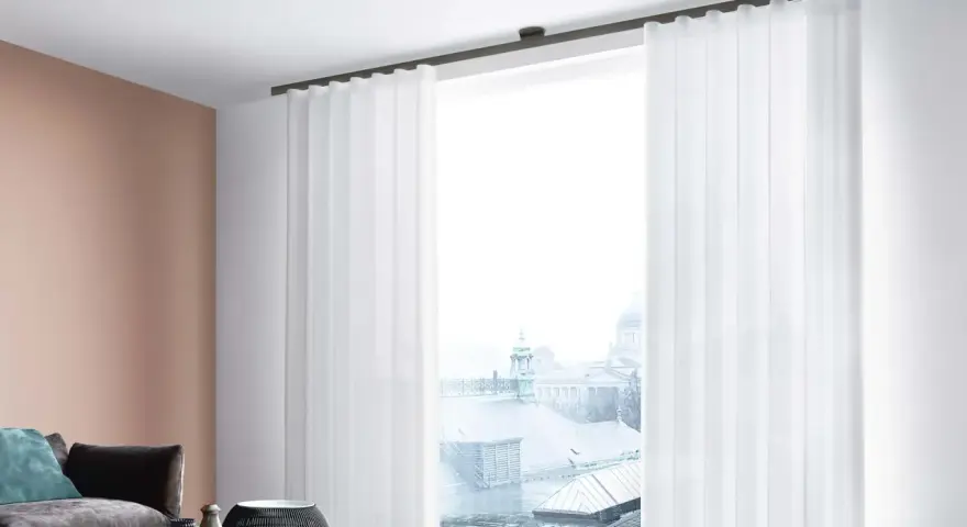 Weiße, transparente Vorhänge mit sanften Wellen und Falten rahmen ein Fenster mit Blick auf eine Stadtkulisse ein.