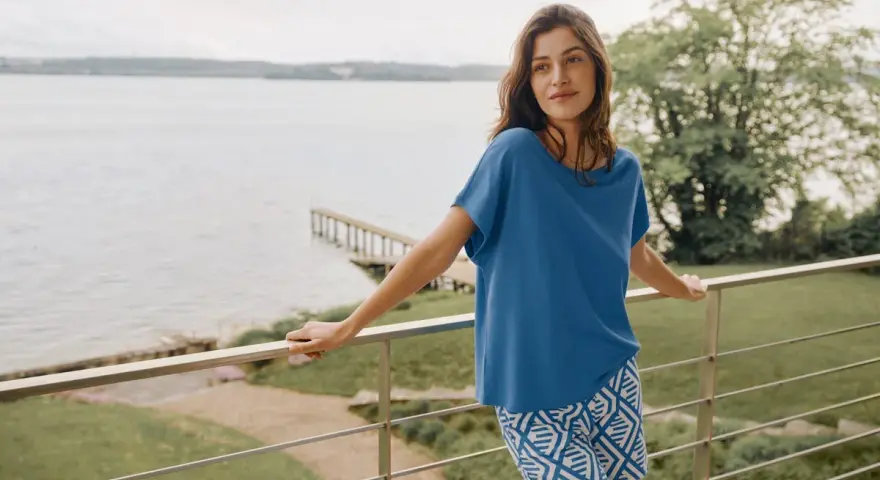 Frau auf Balkon mit Blick auf See, trägt blaues Oberteil und gemusterte Shorts. In Modal schwitzt man nicht, es ist angenehm kühl.