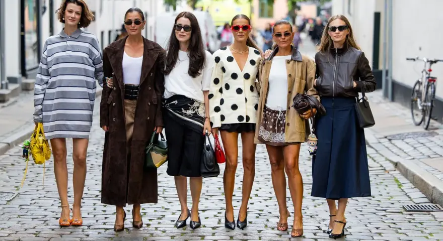 Sechs Frauen in stylischen Outfits, darunter ein gestreiftes Kleid und ein Blazer mit nordischen Mustern, posieren auf einer Kopfsteinpflasterstraße.