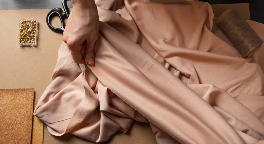 Eine Hand prüft den Fall eines fließenden Stoffes aus Viskose Polyester. Nadeln, Schere und Garn liegen bereit.