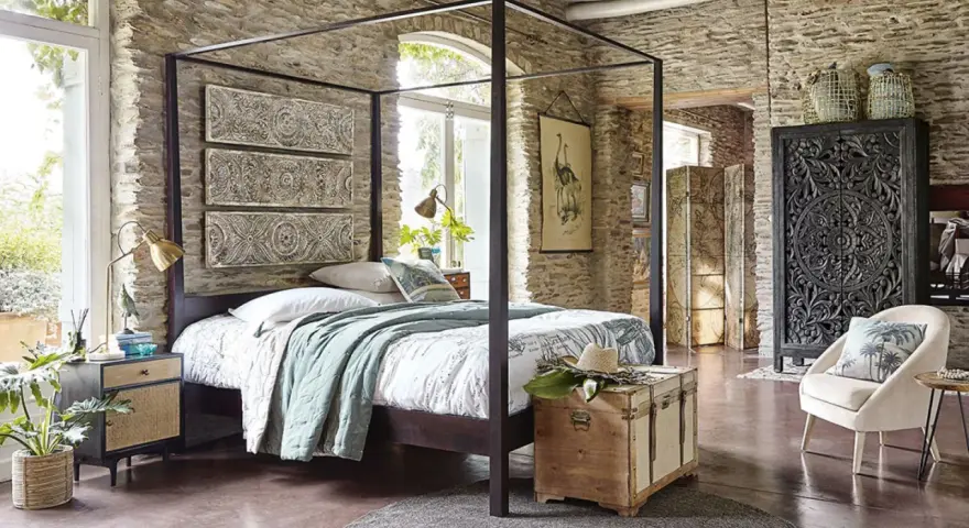 Ein Schlafzimmer mit Steinwänden und einem Himmelbett. Drei Wandpaneele mit orientalischen Mustern hängen über dem Bett. Ein Holztruhe steht am Fußende.