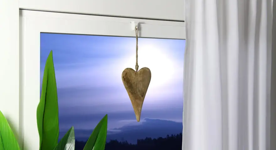 Holzherz hängt an einer Schnur, um die Gardinenstange zu dekorieren. Grüne Blätter und ein dramatischer Himmel im Hintergrund.