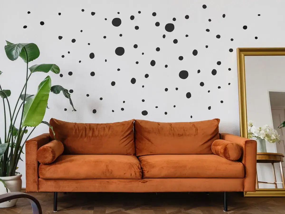 Ein orangefarbenes Samtsofa steht vor einer weißen Wand mit vielen schwarzen Polka Dots. Daneben eine große Pflanze und ein goldener Spiegel.