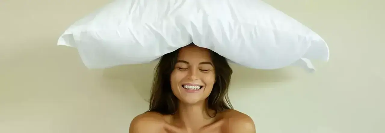 Lachende Frau mit einem weißen Kopfkissen auf dem Kopf, als ob sie es gerade gewaschen hätte und es trocknet.