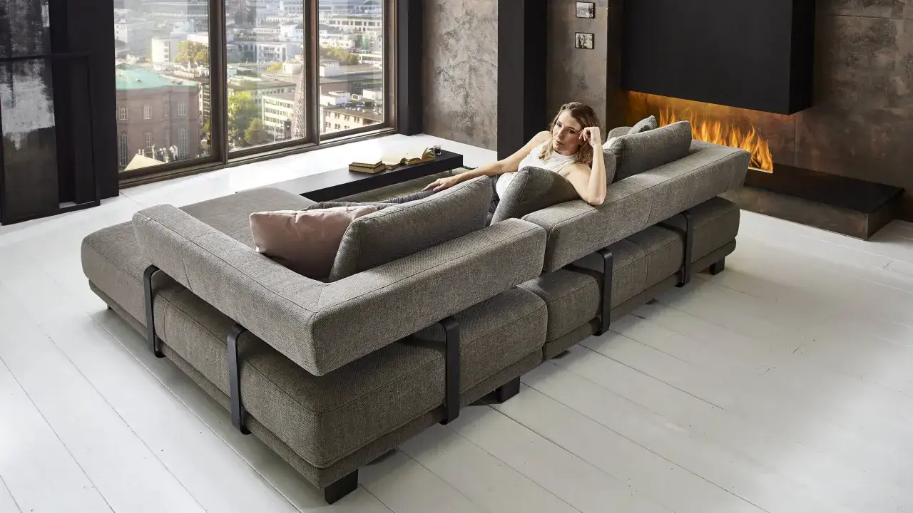 Ein gem&uuml;tliches ecksofa mitten im raum l&auml;dt zum Entspannen ein. Eine Frau liegt darauf, im Hintergrund ein Kaminfeuer.