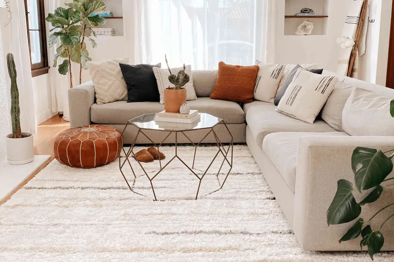 Ein flauschiger, weißer Teppich unter dem Sofa schafft Gemütlichkeit. Darauf ein brauner Pouf und ein moderner Couchtisch.