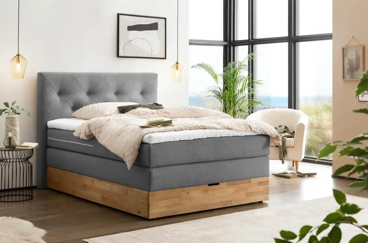 Ein modernes Boxspringbett mit grauem Stoffbezug und Holzsockel steht neben einem großen Fenster mit Meerblick.