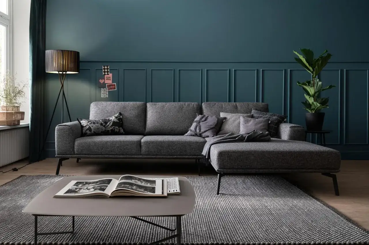 Ein moderner Wohnbereich mit einem anthrazitfarbenen Sofa, das auf einem gemusterten Teppich steht. Kissen und eine Decke zieren das Sofa.