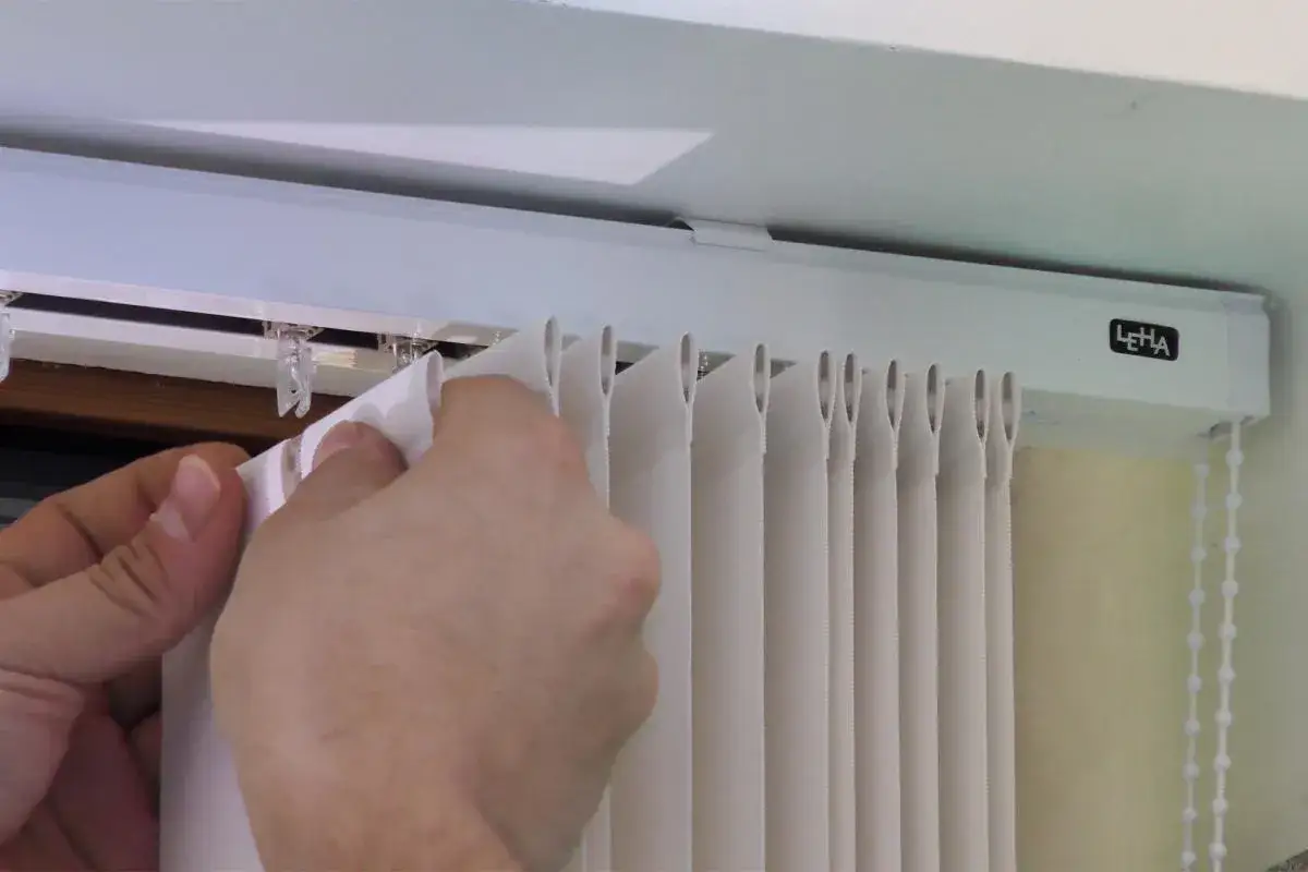 Hände befestigen einen weißen Lamellenvorhang in einer Gardinenschiene. Die Lamellen werden in kleine Clips an der Schiene eingehängt.