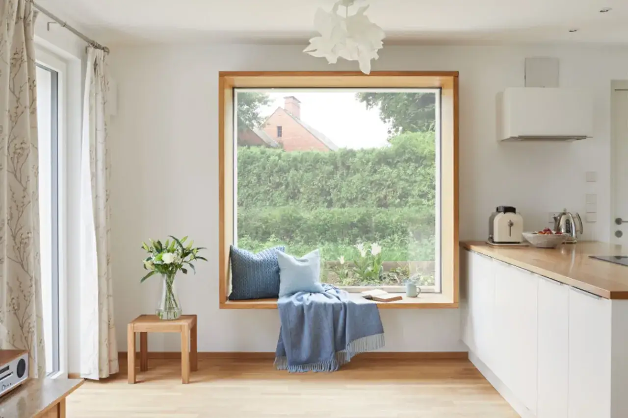 Gemütliche Fenstergestaltung Wohnzimmer ohne Gardinen: breiter Fenstersitz mit Kissen, Decke und Buch, daneben ein kleiner Holztisch mit Blumen.