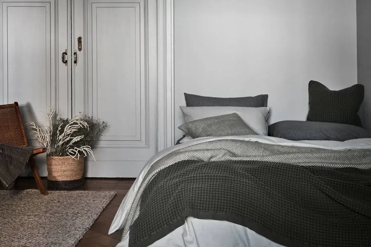Gemütliche Bettwäsche in Grau- und Grüntönen, inspiriert von modernen Bett machen Ideen. Ein Korb mit Trockenblumen und ein Sessel vervollständigen die Szene.
