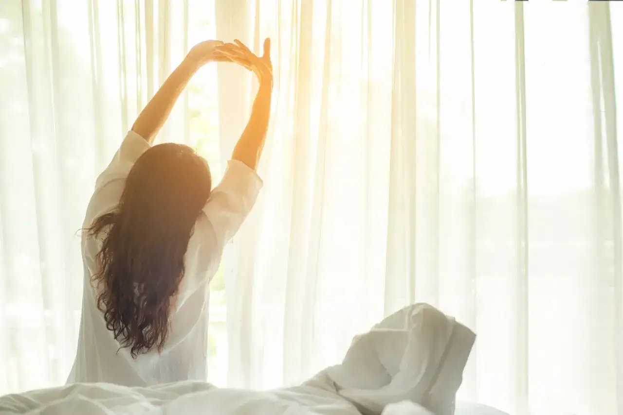 Frau streckt sich im Bett, das Morgenlicht fällt durch die Vorhänge. Wann aufstehen? Die Frage stellt sich bei diesem Anblick nicht.