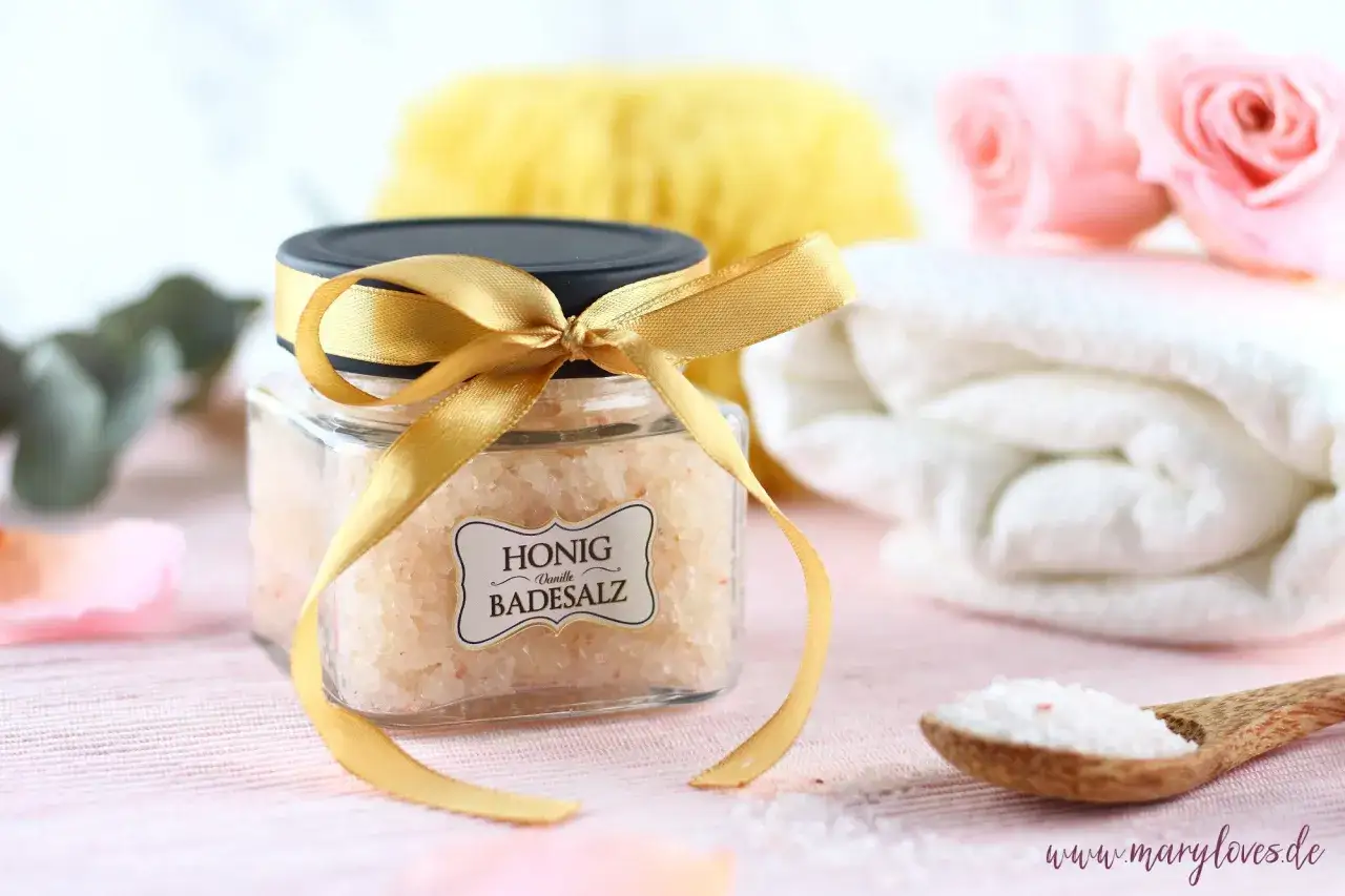 Ein Glas mit selbstgemachtem Honig-Vanille-Badesalz, verziert mit einem goldenen Band. Daneben liegen eine Holzschaufel mit Salz, eine weiche Bürste und gefaltete Handtücher.