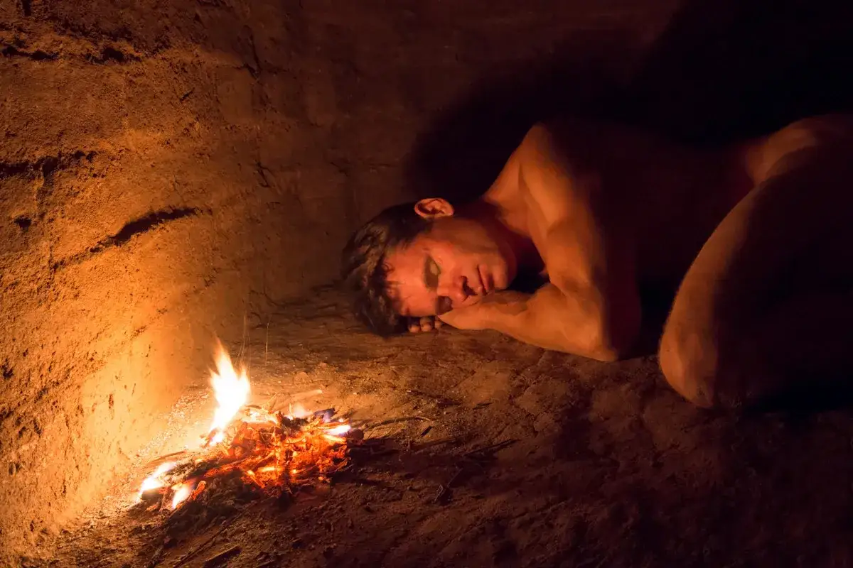Ein Mann schläft neben einem Feuer in einer Höhle. Wer hat das Bett erfunden? Vielleicht dieser Mann, der sich auf dem Boden ausruht.