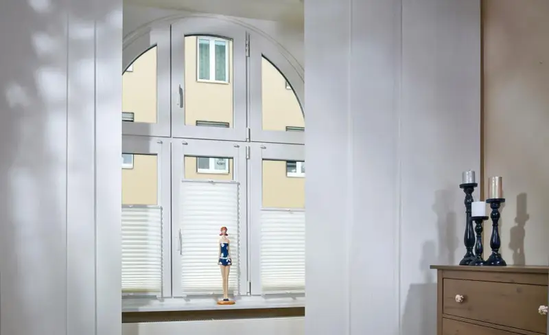 Weiße Schiebegardinen in alten Schienen vor einem Bogenfenster mit Blick auf ein Gebäude. Eine Puppe steht auf der Fensterbank.