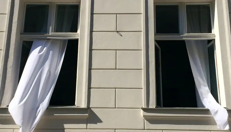 Weiße Gardinen wehen aus zwei offenen Fenstern eines hellen Gebäudes. Die Frage "Gardinen waschen wieviel Grad?" beschäftigt die Hausfrau.